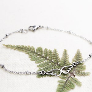 May include: A silver chain bracelet with a delicate wing charm. The bracelet is on a white background with a green fern leaf.