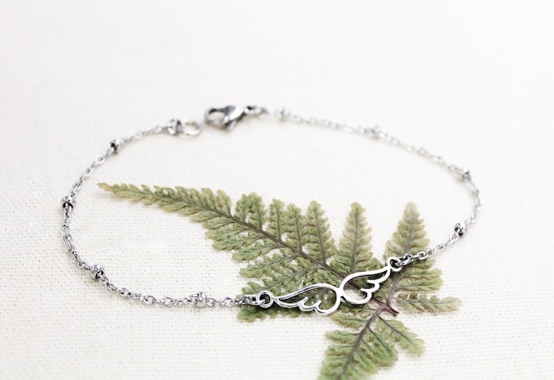 May include: A silver chain bracelet with a delicate wing charm. The bracelet is on a white background with a green fern leaf.