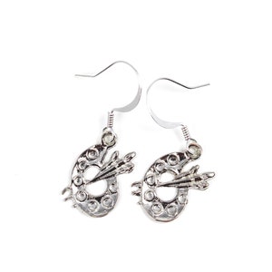 May include: A pair of silver earrings featuring a paint palette design with a paintbrush. The earrings are made of metal and have a simple, elegant design.