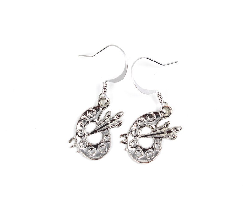 May include: A pair of silver earrings featuring a paint palette design with a paintbrush. The earrings are made of metal and have a simple, elegant design.