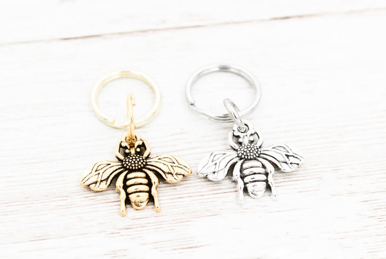 Puede incluir: Dos llaveros con forma de abeja, uno dorado y otro plateado, con un dise&ntilde;o de anillo simple.