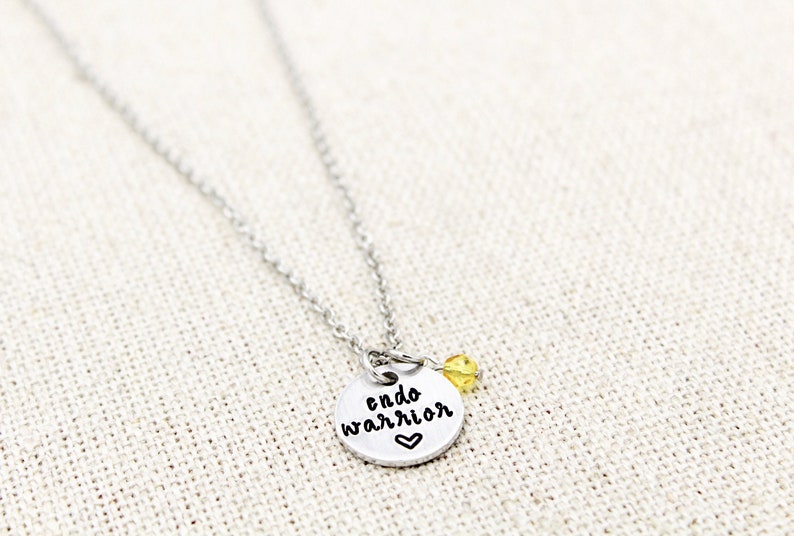 May include: Silver necklace with a round charm that says "endo warrior" with a small heart below the text. A small yellow bead hangs from the charm.