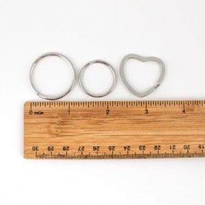 May include: Three silver key rings, one round, one smaller round, and one heart-shaped, are laid out on a wooden ruler with inch markings. The ruler is marked with numbers from 18 to 30.
