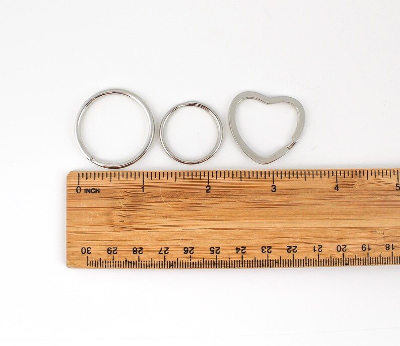 May include: Three silver key rings, one round, one smaller round, and one heart-shaped, are laid out on a wooden ruler with inch markings. The ruler is marked with numbers from 18 to 30.