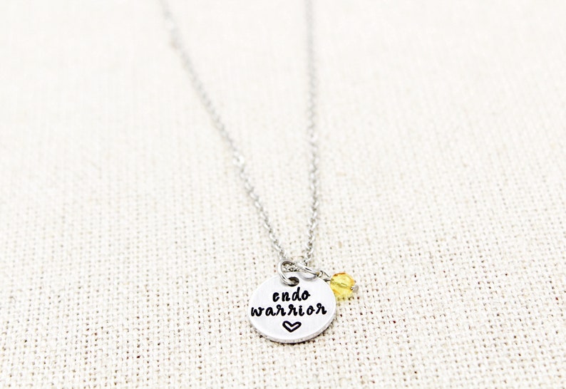 May include: Silver necklace with a round charm that says "endo warrior" with a small heart at the bottom. A small yellow bead hangs from the charm.