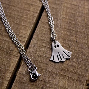 May include: Two silver-colored necklaces with Halloween-themed pendants. One necklace features a ghost pendant, and the other has a small, round pendant. The necklaces are displayed on a rustic, wooden surface.