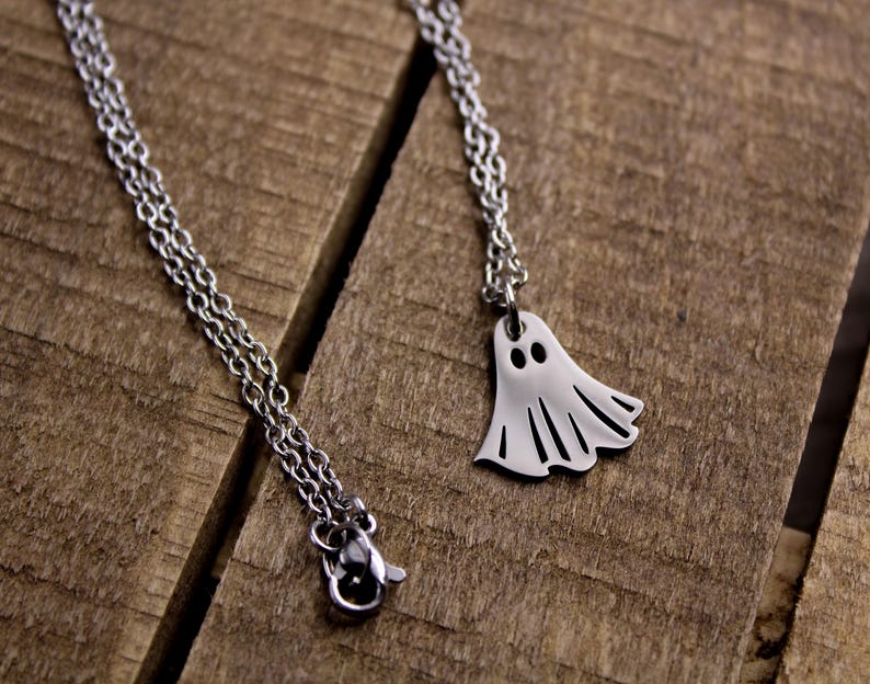 May include: Two silver-colored necklaces with Halloween-themed pendants. One necklace features a ghost pendant, and the other has a small, round pendant. The necklaces are displayed on a rustic, wooden surface.