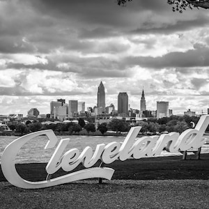 May include: Black and white image of the Cleveland skyline with the city's name in large, white, cursive letters in the foreground. The cityscape includes various buildings and towers under a cloudy sky, with a body of water in the distance.