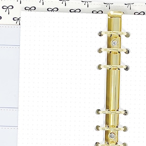 May include: A white six-ring binder featuring a black bow pattern on the interior cover, containing white dot grid paper. A gold-toned metal ring mechanism is attached to the spine. A white pen with gold accents is positioned to the left of the binder.