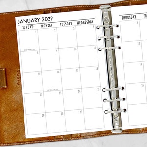 May include: A brown leather-bound planner open to a January 2022 calendar. The calendar pages are white with black text, showing dates and days of the week. The planner is open to a two-page spread.