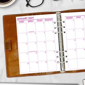 May include: A brown leather Filofax planner open to a January 2022 calendar page. The calendar grid is white with pink text and numbers. The planner is open on a white surface with a coffee cup and glasses in the background.