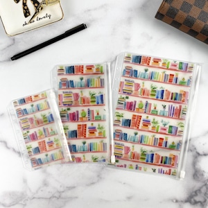 May include: Three clear plastic zipper pouches of varying sizes, each featuring a repeating watercolor-style print of colorful books and small potted plants arranged on shelves. Each pouch includes a white plastic zipper and punched holes for binding.