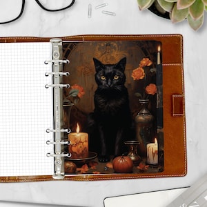 May include: A brown leather planner with a black cat illustration. The planner is open, revealing a grid-lined page and a decorative image of a black cat with candles and flowers. The planner has silver rings.