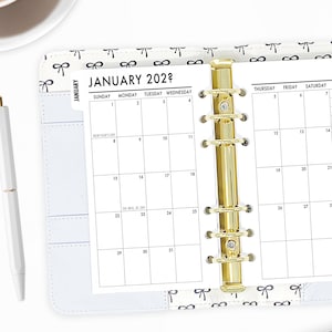 May include: A white planner with a gold coil binding. The planner has a monthly calendar for January 2022 with the days of the week labeled. The planner has a white background with a black and white bow pattern.