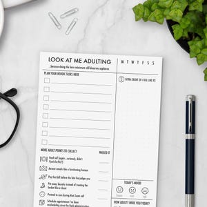 May include: A white notepad with the text "LOOK AT ME ADULTING" and a list of tasks. The notepad includes sections for daily planning, mood tracking, and a checklist of adulting activities. A pen is visible on the right side.