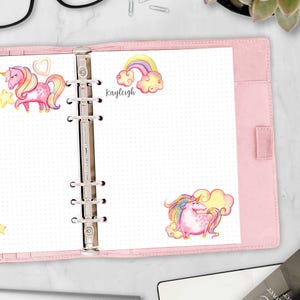 May include: A pink six-ring binder containing dot grid paper. The pages feature watercolor illustrations of pink unicorns, a rainbow with clouds, and a yellow heart. The name Kayleigh is printed in black script near the rainbow illustration.