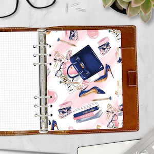 May include: A planner insert with a fashion-themed design. The insert features illustrations of a navy blue handbag, high heels, and accessories on a pink and white background. The planner is open, revealing the insert and a grid-lined page.
