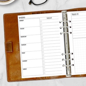 May include: A weekly planner insert with a white background, featuring sections for each day of the week, a to-do list, and a section labeled "Track It!". The planner is held in a brown leather binder.