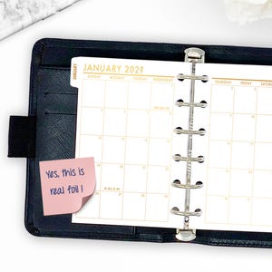 May include: A black leather-like planner open to a January 2022 calendar page. The calendar has gold-colored text and a pink sticky note that reads "Yes, this is real foil!". The planner has a silver binder mechanism.