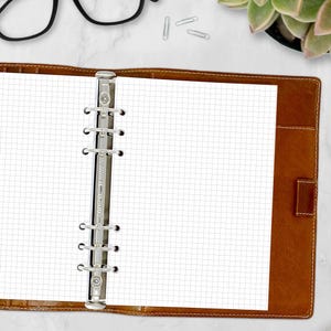 May include: A brown leather binder with a grid-lined paper insert. The binder features a silver metal spine with rings to hold the paper. The paper is white with a light grey grid pattern. The binder is open, revealing the paper.