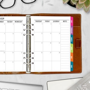 May include: A brown leather bound planner with a silver ring binder. The planner has a monthly calendar view for January 2023 and a weekly view for the first week of January. The planner has color coded tabs for each month of the year, starting with July and ending with June.