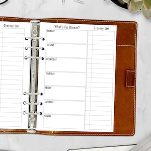 May include: A white planner with a brown leather cover. The planner has a weekly meal planner section with the days of the week listed: Monday, Tuesday, Wednesday, Thursday, Friday, Saturday, Sunday. The planner also has two grocery list sections.