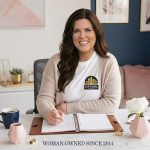 Becki, owner of CityGirl Planners, at her desk in a cream cardigan with the CityGirl Planners logo, planner open in front of her with white flowers nearby. Woman-Owned Since 2014 banner across the bottom. Personal size lined paper planner refills handcrafted by our small team.