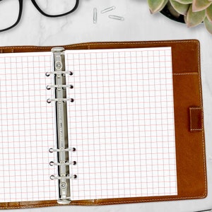 May include: A brown leather bound notebook with a silver ring binder and white gridded pages. The notebook is open to a blank page.