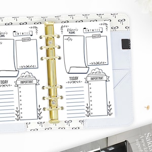 May include: A white planner with black and white floral designs and a gold binder. The planner has sections for "Today", "Important", "Rebecca's Plans", "Notes", "SMTWTFS", and "Gratitude".