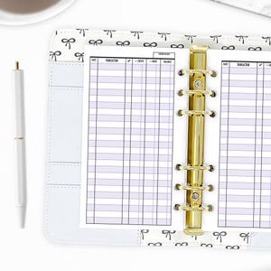 May include: A white binder with a gold ring mechanism and a light purple lined page with columns labeled "Date", "Transaction", "Check", "Credit", "Debit", and "Balance". The binder has a white cover with a repeating pattern of black bows.