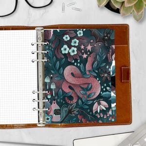 May include: A dark blue planner with a pink snake, flowers, and stars on the cover. The planner has a silver ring binder and the text "GOTI" is visible on the cover.