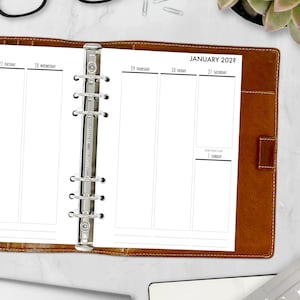 May include: A brown leather bound planner with a silver ring binder. The planner is open to the January 2022 calendar page. The calendar is divided into daily columns with the days of the week labeled at the top. The calendar is blank, ready for writing.
