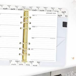 May include: A white planner with a gold binder ring system. The planner has a black and white bow pattern on the cover and the text "JANUARY 2023" printed on the front. The planner is open to the first page of the month, which has the days of the week listed and space for writing.
