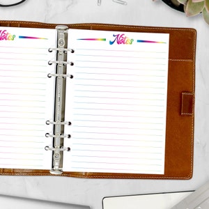 May include: A brown leather binder with lined paper inserts. The paper has a rainbow-colored header with the word "Notes" and rainbow-colored lines. The binder is open, revealing the paper and metal rings.