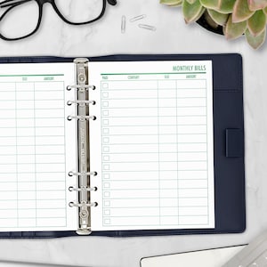 May include: A navy blue binder open to a monthly bills planner page. The page has green grid lines and the words "MONTHLY BILLS" at the top. The binder is open, revealing the metal rings and the words "Time-Catcher" on the spine.
