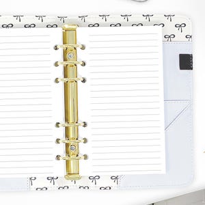 May include: A white, lined,  paper refill for a  gold-toned, six-ring binder. The binder has a white cover with a black and white bow pattern.