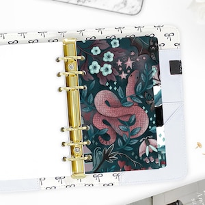 May include: Open white planner with gold rings, featuring a decorative insert with a pink snake illustration on a dark teal background. The insert also includes floral and celestial elements.