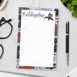 May include: A notepad with a purple border featuring black cat illustrations and the text "#witchyplans". A cartoon bat graphic is above the text. A black pen and glasses are next to the notepad. The notepad is blank and ready for notes.