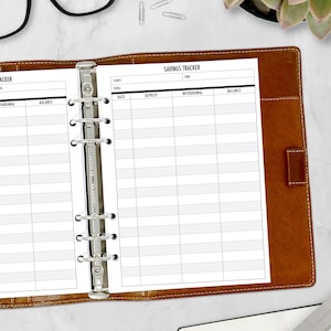 May include: A white page with a black grid and the text "Savings Tracker" at the top. The page is in a brown leather binder with silver rings.