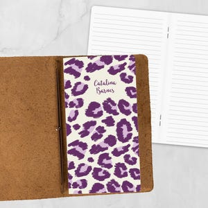 May include: A personalized journal with a purple leopard print cover and the name "Catalina Barnes" in script. The journal is open, revealing lined pages, and is encased in a brown leather cover.