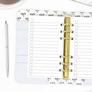 May include: A white planner with a gold binder and lined paper. The planner has a bow pattern on the cover and a matching white pen. The planner is open to a to-do list page.