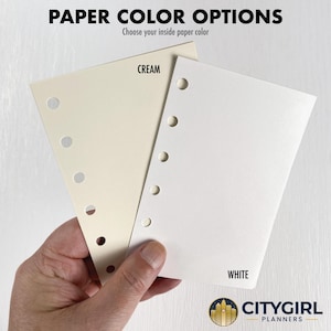 Paper color options guide showing a hand holding two punched paper samples — natural cream on the left, bright white on the right — against a light wood background. Both are 80 pound text weight, buttery smooth, and write with most pens and pencils without bleed-through. Not recommended for alcohol-based markers. Choose your inside paper color at checkout. CityGirl Planners logo displayed at bottom right.