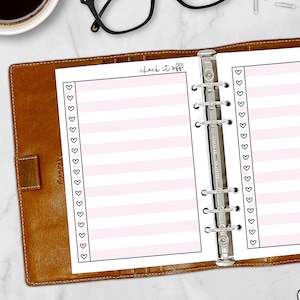 May include: A planner insert with pink striped lines and a row of black heart icons. The text "check it off!" is written in a handwritten style. The insert is held in a brown leather binder with silver rings.