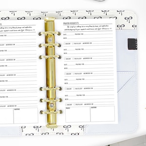 May include: A white prayer journal with a gold binder and a black and white bow pattern on the cover. The journal has a section for prayer requests with space to note the date, need, if it is urgent, if it has been fulfilled, and when it was answered. The journal also has a section for praises and gratitude.