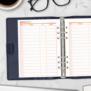 May include: A navy blue binder with a "MONTHLY BILLS" insert. The insert has orange lines and text for tracking payments. A coffee cup, glasses, and paper clips are in the background.