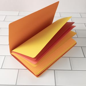 May include: An open notebook with a hard orange cover and a fan of colorful pages. The pages are in shades of yellow, orange, and red, with a dotted pattern. The notebook is on a white tiled surface.