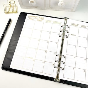 May include: A black leather binder open to a calendar page for January 2026. The calendar grid is printed in gold, with the days of the week across the top. A black pen and gold paper clip are visible in the background.
