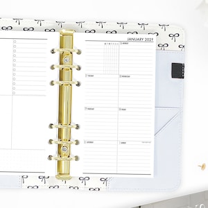 May include: A white planner with a gold binder ring system. The planner has a weekly layout with a section for each day of the week. The cover of the planner has a pattern of black bows on a white background. The planner is open to the week of January 26th to February 1st, 2029.