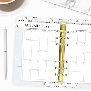 May include: A white planner with a gold binder and a January 202? calendar page. The planner has a bow pattern on the cover and a white pen is on the left side. The calendar grid is black and white.
