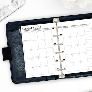 May include: A black leather planner open to a January 2022 calendar page. The calendar is white with black text, showing the days of the week and dates. The planner has a silver binder mechanism and a black elastic closure.